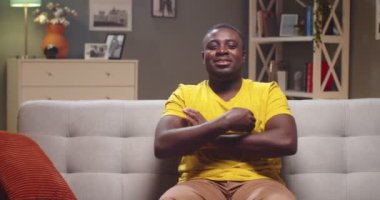 Genç Afro Amerikalı adam oturma odasında modern iç tasarımı ile kanepede dinleniyor. Yakışıklı adam gülümsüyor, kameraya bakıyor ve evde kollarını kavuşturuyor. Rahatla, rahatla, izolasyon kavramı.