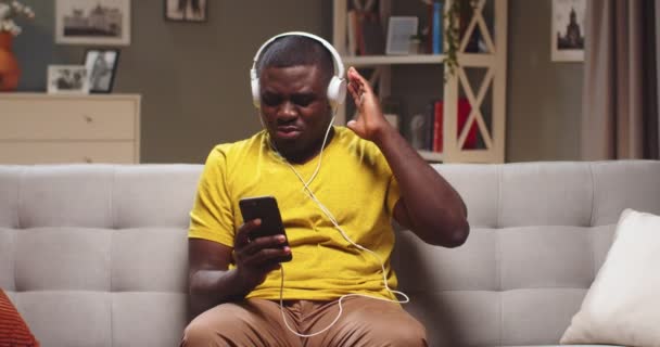 Portret wesołego mężczyzny w słuchawkach słuchającego muzyki i chłodzącego się wieczorem na kanapie. Młody Afroamerykanin tańczący, trzęsący głową i trzymający smartfona w domu. Koncepcja rozrywki. — Wideo stockowe