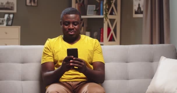 Ung afroamerikan sitter på soffan och vilar och använder smartphone. Snygg kille som sms:ar vänner, tittar på videor och bläddrar bland nyheterna på kvällen. Internet, begreppet sociala medier. — Stockvideo
