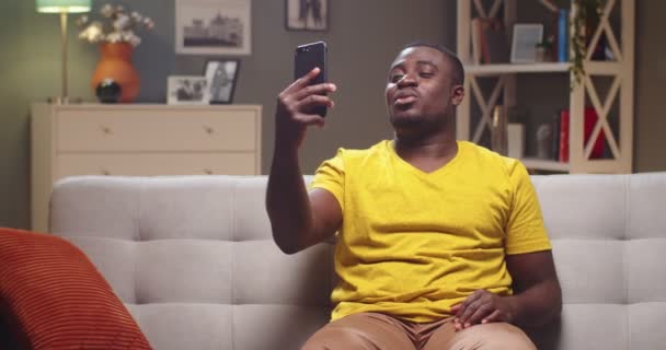 Encantador hombre sentado en el sofá y llamando a sus amigos en casa por la noche. Jóvenes afroamericanos hablando por teléfono vía videoconferencia durante la cuarentena. Aislamiento, concepto de comunicación. — Vídeos de Stock