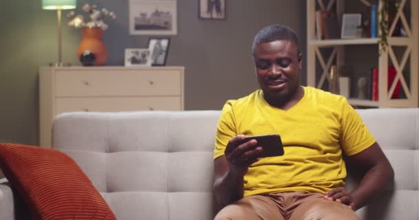 Hezký mladík sedí na gauči a večer se dívá na video na smartphonu. Afroameričan odpočívá a odpočívá doma v obývacím pokoji. Zábava, technologický koncept. — Stock video