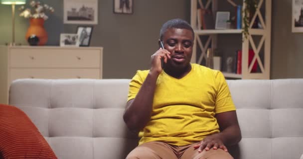 Yakışıklı bir adam kanepede dinleniyor ve oturma odasında sohbet ediyor. Yetişkin Afrikalı Amerikalı, akşam işten sonra arkadaşlarıyla konuşuyor. Teknolojiler, iletişim. — Stok video