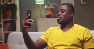 Açık fikirli bir adamın, arkadaşını arayıp akşam telefonda konuştuğu portresi. Yetişkin Afro-Amerikalı adam kanepede oturuyor ve evde video konferansı yapıyor. Sosyal medya, iletişim konsepti.