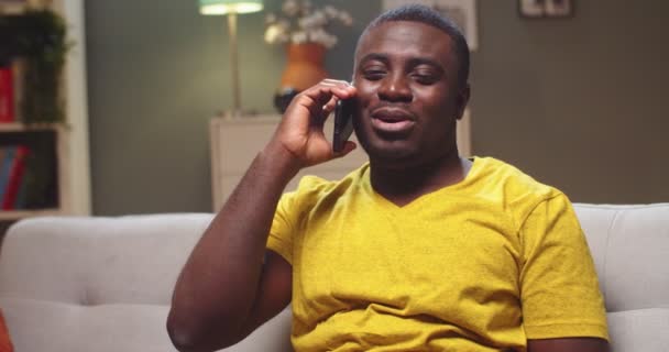 Vidám férfi afro-amerikai lazulás a kanapén a nappaliban este. Fiatal fickó hívja a barátját, telefonon beszél és nevet. Kommunikáció, technológiák és eszközök fogalma. — Stock videók