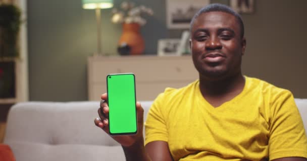 Портрет красавчика, який сидить на дивані і тримає смартфон з зеленим екраном. Молодий афроамериканець показує хроматичний ключ до камери і посміхається. Вертикальна позиція. Домашня концепція. — стокове відео