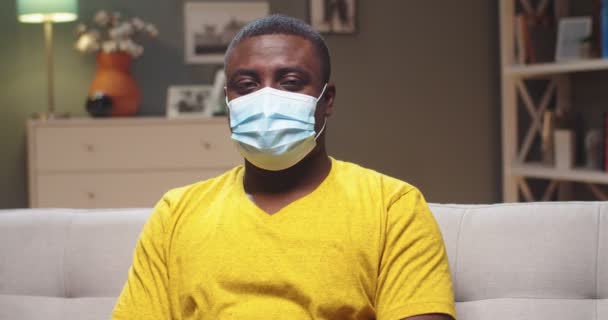 Porträtt av charmig man i medicinsk skyddsmask vilar på soffan i vardagsrummet. Vuxen man afro amerikan ataying hem och tittar på kameran. Begreppet pandemi, coronavirus och karantän. — Stockvideo