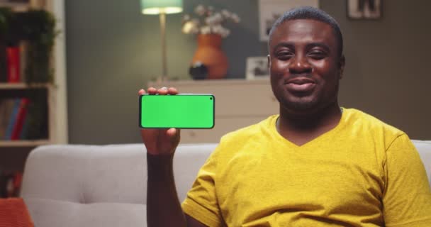 Gut aussehender Mann, der ein digitales Smartphone mit Chroma-Schlüssel in der Hand hält und abends lächelt. Afroamerikanischer Typ mit grünem Bildschirm und Blick in die Kamera zu Hause. Horizontale Position. — Stockvideo