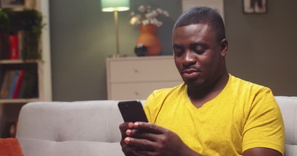 Porträtt av snygg kille vilar efter hård arbetsdag i vardagsrummet hemma. Ung afro amerikansk man messaging vänner, skriva på smartphone och leende. Sociala medier, teknikkoncept. — Stockvideo