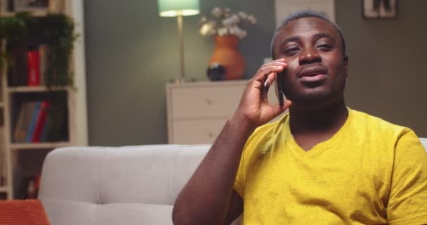 Portrét okouzlujícího Afroameričana, jak volá svému příteli a večer mu říká dobré zprávy. Mladý muž sedící na gauči a telefonující doma. Komunikace, technologie, koncepce připojení. — Stock video