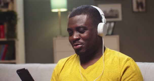 Porträtt av attraktiv kille chilling på soffan och lyssna på musik i vardagsrummet. Ung hane afro amerikan i hörlurar med smartphone, skakar huvud för att slå. Teknik, underhållning. — Stockvideo