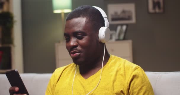 Retrato del hombre relajado descansando en el sofá, escuchando música por la noche en casa. Hombre afroamericano adulto disfrutando de la canción, sacudiendo la cabeza y el uso de teléfono inteligente en la sala de estar. Tiempo libre, entretenimiento. — Vídeo de stock