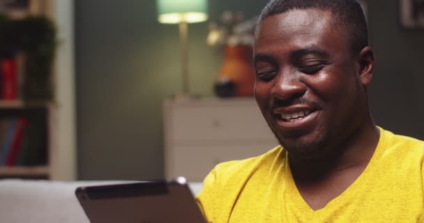 Charmanter Mann, der abends auf der Couch chillt und zu Hause ein digitales Tablet in der Hand hält. Junger Afroamerikaner schaut Komödienfilm und lacht im Wohnzimmer. Unterhaltung, Internetkonzept. — Stockvideo
