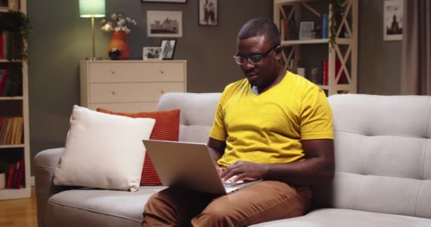 Chytrý mladík sedí na gauči a večer píše na notebook. Afroameričan v brýlích píše knihu a píše akademické noviny v obývacím pokoji doma. Vzdělání, koncepce copywriteru. — Stock video