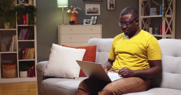 Поганий хлопець у окулярах сидить на дивані і ввечері використовує ноутбук удома. Дорослі афроамериканці серфінгують Інтернет, відпочиваючи після важкого робочого дня. Технології, соціальні медіа, розваги. — стокове відео
