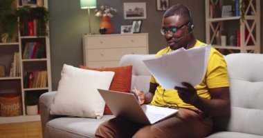 Tecrübeli destek uzmanı belgeleri kontrol ediyor ve internetten danışmanlık yapıyor. Akşamları karantina sırasında evde çalışan kulaklıklı yetişkin Afro Amerikalı bir adam. Covid-19, salgın, telgraf.