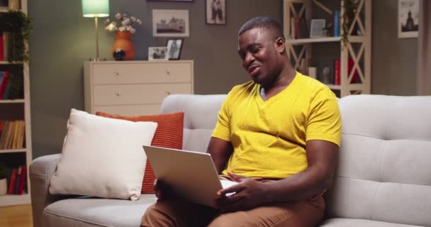 Attraktiver junger Mann, der auf der Couch sitzt und mit der Familie spricht, Freundin abends am Laptop. Afroamerikaner bei Videokonferenzen, Verabschiedung und Küsschen zu Hause. Kommunikationskonzept. — Stockvideo