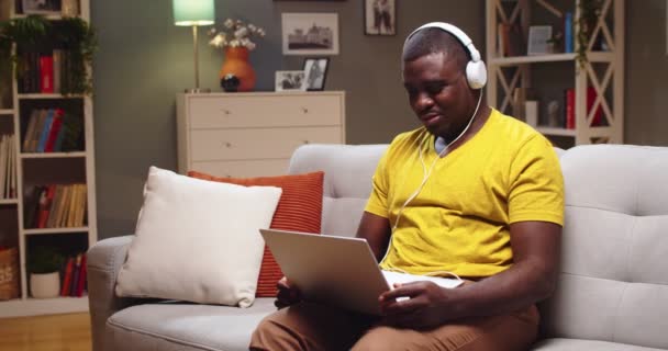 Hombre guapo en auriculares escalofriantes en el sofá y el uso de la computadora portátil en la noche. Joven afroamericano escuchando música, bailando y trabajando en la computadora en casa. Concepto de entretenimiento. — Vídeos de Stock