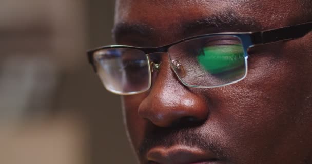 Atractivo chico afroamericano sentado en el sofá, trabajando y escribiendo en el portátil. Primer plano de la cara del hombre en gafas con reflejo de luz verde en la noche. Remore trabajo, negocio, trabajo, concepto de educación. — Vídeos de Stock