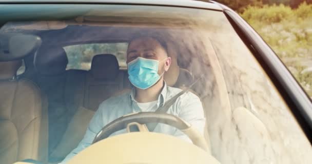 Primer plano del joven conductor guapo con máscara azul médica a caballo coche y escuchar música, moviendo las manos en rima. Cielo reflejado en las ventanas del coche. Estilo de vida, carretera, coche, concepto de conductor. — Vídeos de Stock