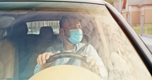 Menutup pengemudi muda tampan dengan masker biru medis naik mobil dan mendengarkan musik, bergerak tangan dalam ritme dan melambaikan tangan kepada orang-orang di luar. Langit tercermin dalam jendela mobil. — Stok Video