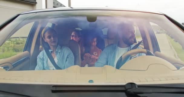 Front view of parents and excited doughters smiling driving car in city. Young family waving to new neighbors after successful home purchase. Concept of lifestyle, happy family, travel together. — Stock Video