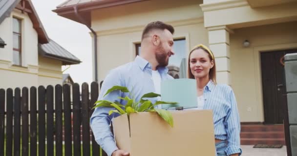 Hermosa pareja joven esperando a que el coche se mueva, la celebración de la caja con flores y lámpara en el fondo nueva casa. La familia se muda a un nuevo apartamento en una zona encantadora en un día soleado. Concepto de estilo de vida, familia feliz. — Vídeo de stock