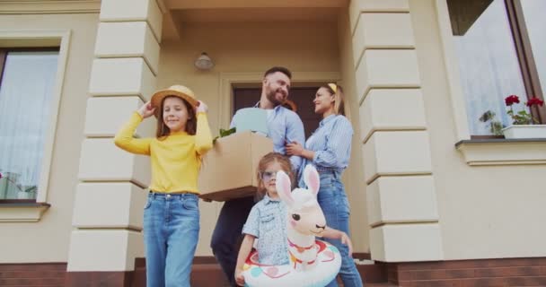 Maravilhosa adorável família jovem alegre com crianças ficar na frente da nova casa e segurando caixa de papelão com plantas durante a mudança para novo apartamento. Belos donuts a divertirem-se. Menina vestindo cabana. — Vídeo de Stock