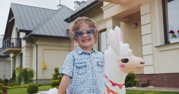 可爱的小女孩玩肥皂泡，戴着太阳镜站在一个漂亮的院子里，一个舒适漂亮的房子里，有一个美洲驼形状的救生圈。生活方式、幸福家庭的概念. — 图库视频影像
