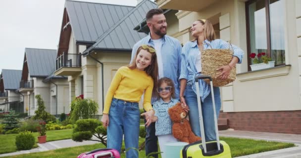 Lycklig familj med saker redo att flytta till ett annat land stående med resväskor mot bakgrund av deras gamla sålda hus och leende tittar på kameran. Familjen förbereder sig för resan. — Stockvideo
