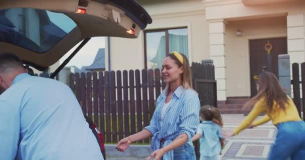 Leuke familie stapt uit de auto en laadt dozen uit naar het nieuwe huis terwijl de kinderen spelen. Ouders verhuizen naar een nieuw appartement in een mooie omgeving op een zonnige dag. Concept van levensstijl, gelukkig gezin. — Stockvideo