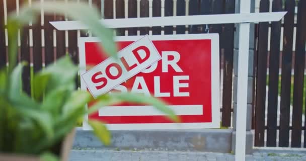 Young couple standing in front of sold sign looking at newly purchased home. Family moving to a new apartment area at sunny day. Husband holding box with plant wife showing keys to new home. — Stock Video