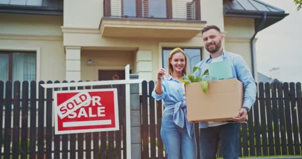 Beautiful young couple staying holding box with flowers and lamp at background new house and sold sign. Wife showing keys to new apartment lovely area on sunny day. Concept of lifestyle, happy family. — Stock Video