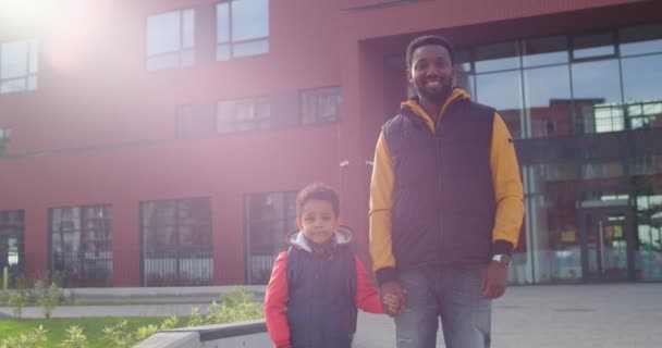 Fröhliche junge afroamerikanische männliche Eltern, die mit ihrem niedlichen kleinen Sohn im Freien stehen. Porträt eines glücklichen Vaters mit einem Erstklässler in der Nähe der Schule, der in die Kamera lächelt. Herbstkonzept — Stockvideo