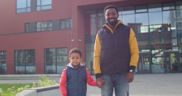 一个快乐的非裔美国人与可爱的小儿子小学生一起站在校园前的画像。年轻的爸爸带着一年级的学生去学校，微笑着。教育概念 — 图库视频影像