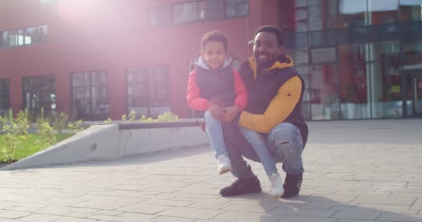 Радісний молодий батько, що стоїть з милим маленьким сином-молодшим студентом на вулиці в доброму настрої в сонячний ранок. Щасливий афроамериканець з малою ученицею біля школи посміхається до фотоапарата — стокове відео