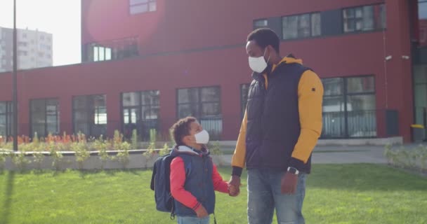 Retrato de un hombre afroamericano feliz y un lindo colegial con máscaras en el patio de la escuela en un día soleado. Papá joven con alumno de primer año mirando a la cámara en la calle al aire libre. Concepto escolar — Vídeos de Stock