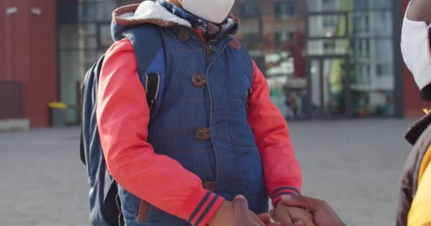 Gros plan d'un petit écolier afro-américain mignon masqué debout avec un sac à dos et parlant avec son père à l'extérieur. Jeune homme parlant et remonter le moral fils mignon le premier jour de l'école. Concept de famille — Video