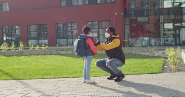 Όμορφος άντρας με μάσκα στέκεται στο δρόμο κοντά στο σχολείο και προετοιμάζει το χαριτωμένο αγοράκι για την πρώτη σχολική μέρα. Αφροαμερικανός πατέρας και ο γιος του - μαθητής δημοτικού σχολείου στο δρόμο στην πόλη — Αρχείο Βίντεο