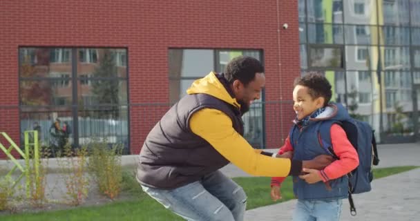 Porträtt av glad ung man på gott humör promenader i stan med liten skolpojke efter skolan på solig dag. Lycklig far tar med sig en liten elev med ryggsäck på händerna utomhus. Familjeförhållanden — Stockvideo