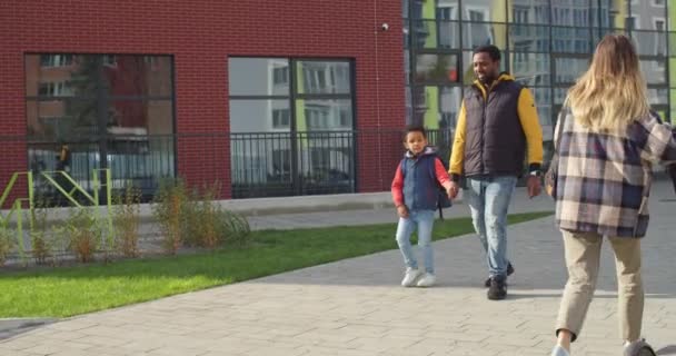 Neşeli genç baba, güneşli bir sabahta okula giden küçük okul çocuğuyla sokakta yürüyor. Babasıyla birlikte sırt çantalı küçük bir öğrenci. Aile ilişkileri. Eğitim kavramı — Stok video