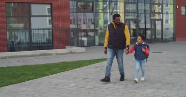 Joven feliz afroamericano padre varón caminando en la calle con su pequeño hijo lindo de la escuela en el día soleado. Guapo padre hablando con junior shool estudiante con mochila después de lecciones al aire libre. — Vídeos de Stock