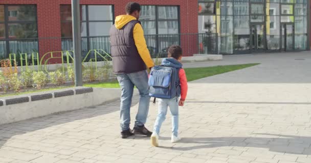 Rear of African American man walking with little son schoolboy to school. Loving father brings first year pupil with backpack to lessons and boy running to school on sunny morning. Education concept — Stock Video