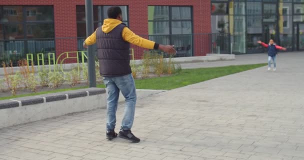 Im Hintergrund ein afroamerikanischer fürsorglicher, liebevoller Vater, der seinen kleinen lächelnden, fröhlichen Sohn aus der Schule auf der Straße trifft. Glückliches afroamerikanisches Schüler-Kind, das gut gelaunt nach dem Unterricht zum Vater rennt. Familienkonzept — Stockvideo