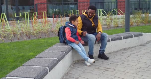 Feliz guapo afroamericano amoroso padre varón sentado en el banco cerca de la escuela con su pequeño hijo alumno y darle paquete con el almuerzo antes de clases. Alegre colegial con mochila corriendo a la escuela — Vídeos de Stock