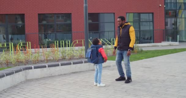 Счастливый красивый афроамериканец отец и сын стоят на улице рядом со школой и болтают в хорошем настроении. Радостный школьник говорит с отцом и показывает ему что-то. Возвращение в школу — стоковое видео