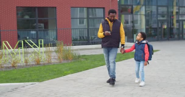 Unga afroamerikanska manliga förälder promenader på gatan i staden med sin lilla son från skolan och skriva på smartphone. Stilig far bläddrar och sms: ar på mobilen medan du går med barn — Stockvideo