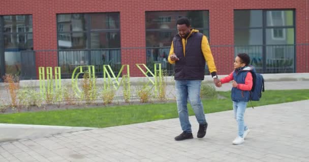 Tânărul tată afro-american fericit mergând pe stradă cu fiul său mai mic după școală și tastând pe smartphone. Tată fericit atingând și mesaje text pe telefonul mobil în timp ce mergeți cu copilul și discutați — Videoclip de stoc