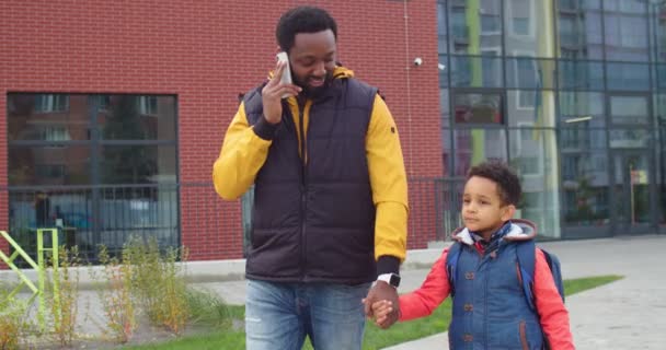 Портрет афроамериканского любящего отца, идущего с маленьким сыном из школы и звонящего по смартфону на улице. Счастливый мужчина разговаривает по мобильному телефону, когда идет с маленьким школьником. Семейная концепция — стоковое видео