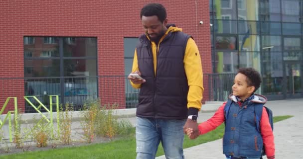 Afroamericano padre amoroso caminando con su pequeño hijo de la escuela y escribiendo en el teléfono inteligente al aire libre. Mensajes masculinos alegres en el teléfono móvil mientras que va con pequeño colegial. Concepto familiar — Vídeo de stock