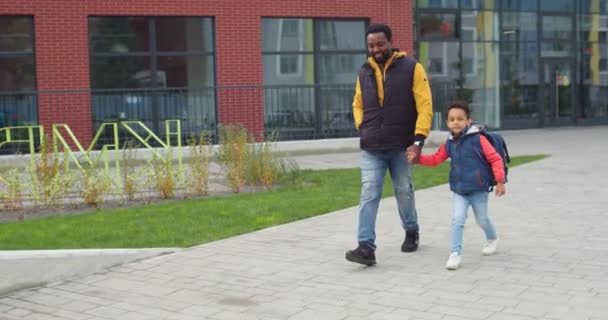 Pequeño niño afroamericano con mochila y su padre tomados de la mano mientras caminaba desde la escuela. Alegre papá sonriente y niño caminando y saltando en la calle. Concepto de educación infantil — Vídeo de stock